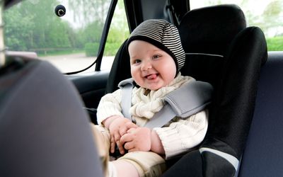 婴儿戴着帽子，穿着毛衣，坐在汽车座椅上微笑。