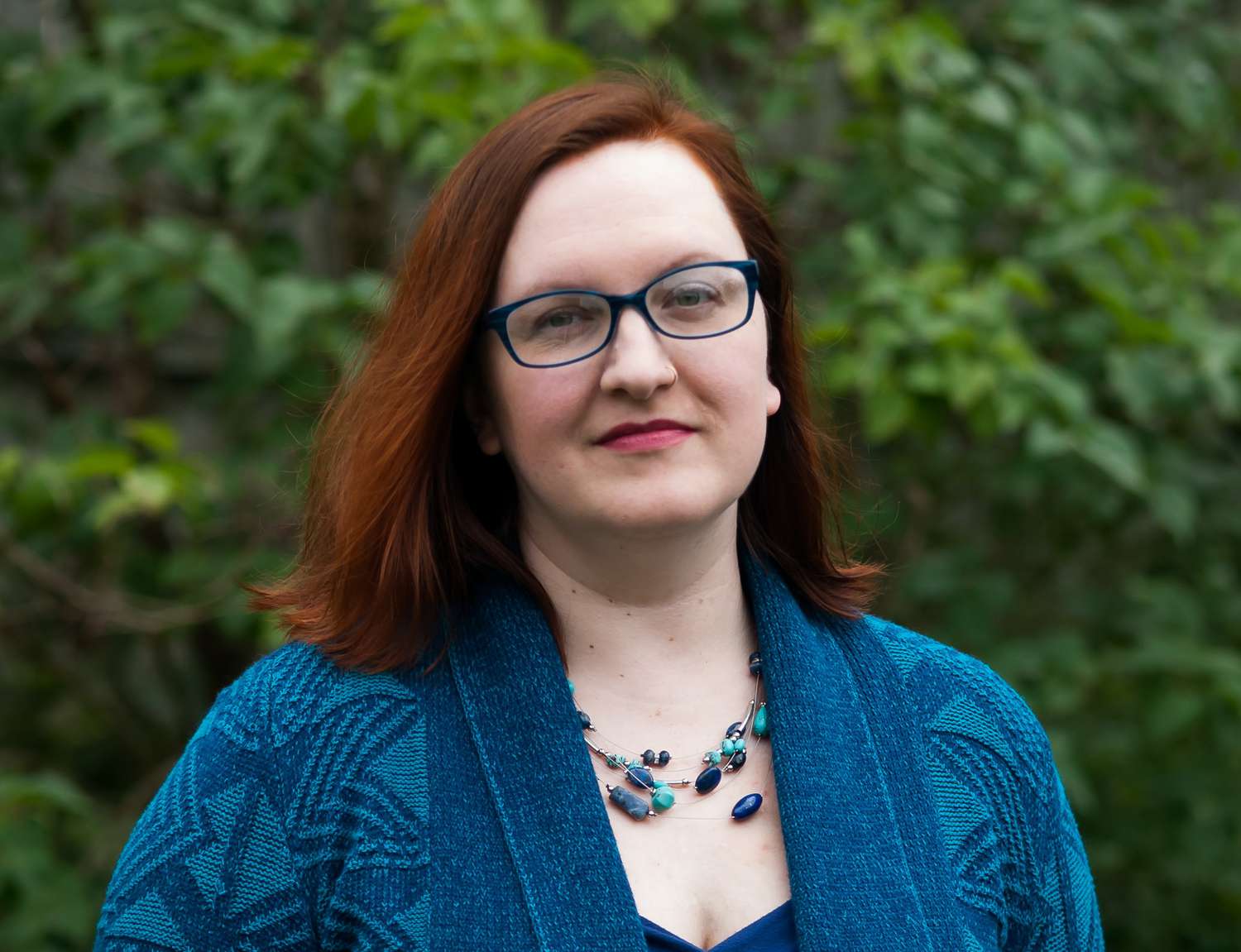 A white woman with red hair and blue glasses stands in front of green trees