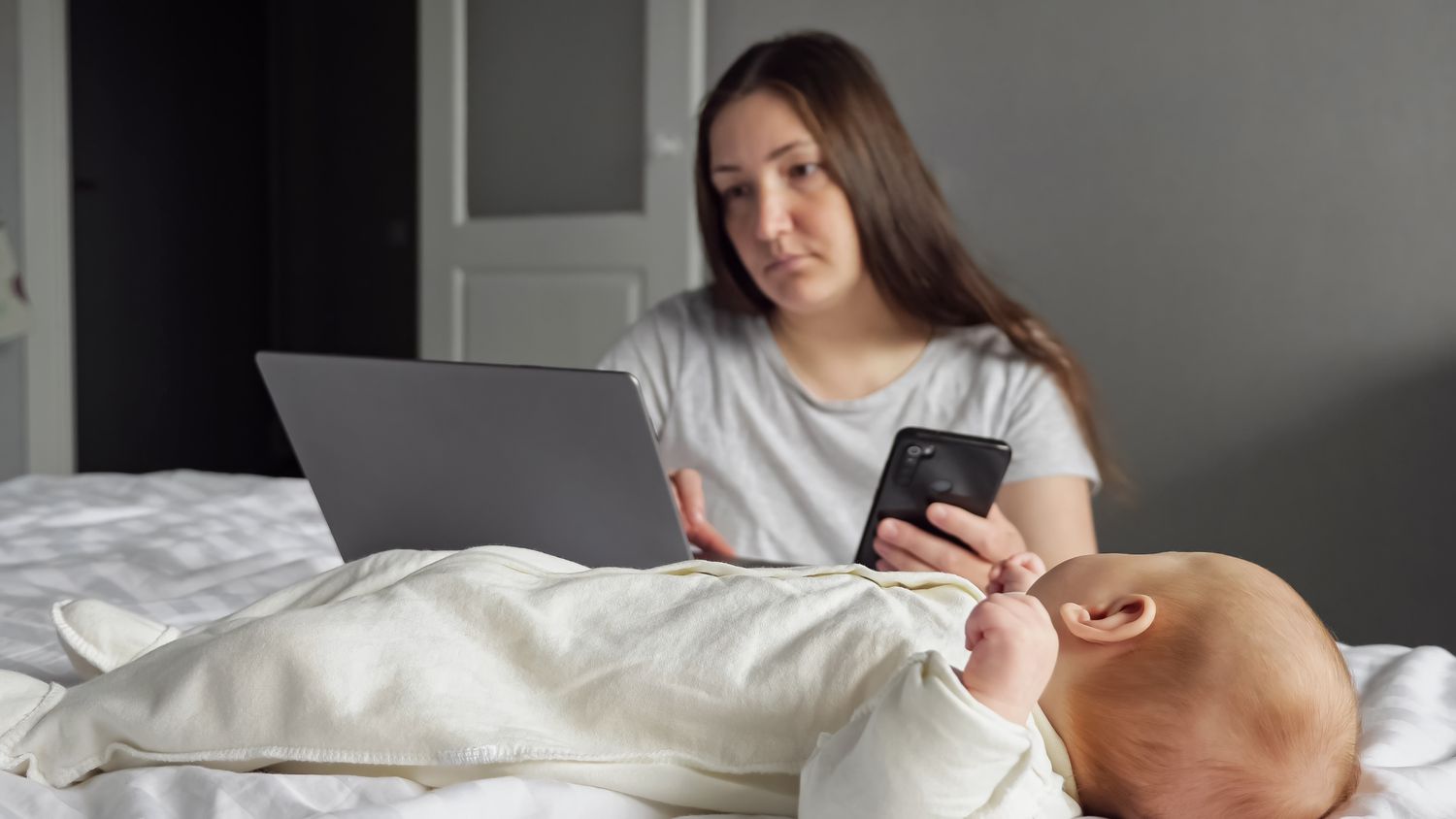 Parent looks at phone and computer while infant is lying on bed