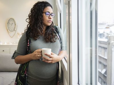 pregnant woman looking out window drinking coffee