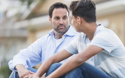 A father sitting with his teenage son on the curb in front of their house.