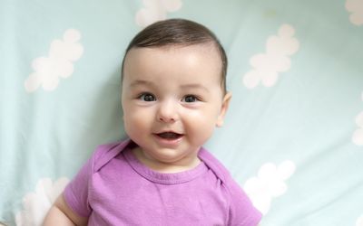 Baby boy in purple onesie looking at the camera and smiling lying against cloud sheets