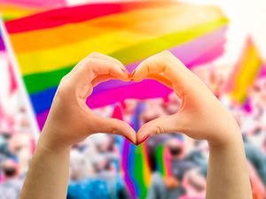 在夏季同性恋骄傲游行场外飘扬的彩虹旗前，支持性的手做心形手势并挥手