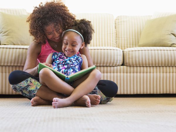 Mixed race mother and daughter reading book on floor near sofa